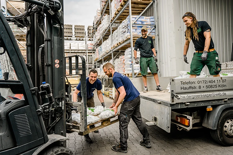 Zwei Mitarbeiter von Mobauplus stehen auf der Ladefläche eines Pritschenwagens und helfen gemeinsam mit zwei Handwerkskunden bei der Beladung.